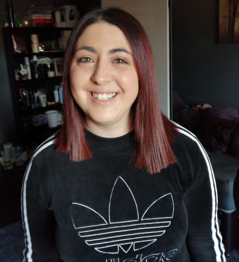 A photo of Hannah smiling, she has purple shoulder length hair and is wearing an adidas jumper.